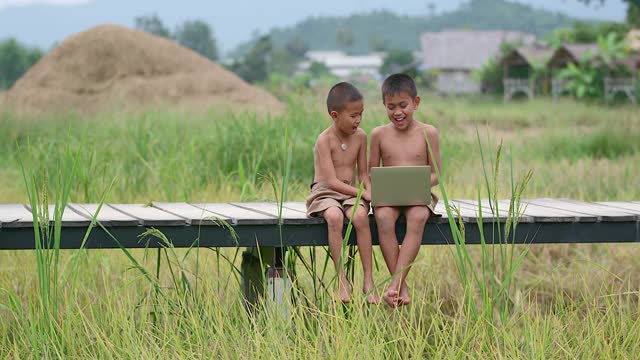 两个农村男孩愉快地玩着笔记本。在笑脸上在木桥上以田野为背景的接入方式，技术的到来在荒野中仍然可以使用互联网。视频素材
