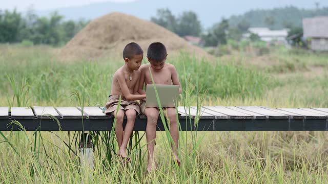 两个农村男孩愉快地玩着笔记本。在笑脸上在木桥上以田野为背景的接入方式，技术的到来在荒野中仍然可以使用互联网。视频下载
