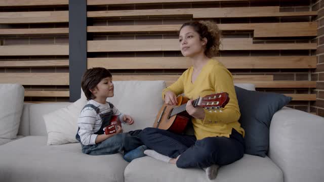 美丽的年轻妈妈弹吉他和她的儿子都有乐趣的微笑和唱歌视频下载