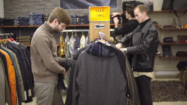 年轻的白人男人在黑色星期五大减价时在商店里挑选衣服。三个集中的英俊男性顾客在购物中心挑选服装。折扣与经营理念。视频下载