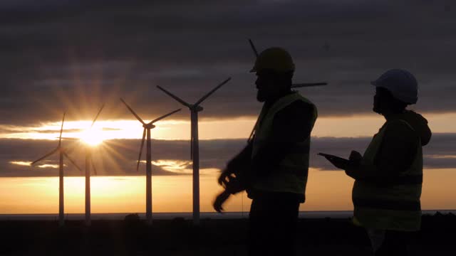 可再生能源系统。黄昏时分，电力维护工程师在风力涡轮机发电站附近的田里工作，身后是阴沉的天空。两位工程师同事的商务会议。团队合作。视频素材