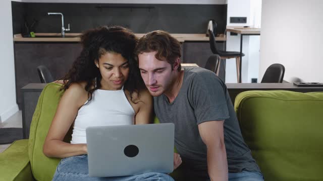快乐的多种族年轻夫妇使用笔记本电脑一起在网上购物，坐在客厅的沙发上看视频电影。微笑的白人男子和非洲裔美国妇女在家里看电脑。视频素材