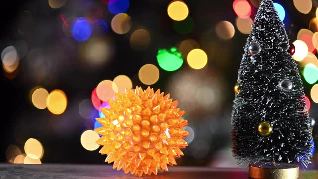 一个黄色的尖球，可能代表一种冠状病毒，靠近一棵微型圣诞树。圣诞彩灯的背景。潘相机运动。视频素材