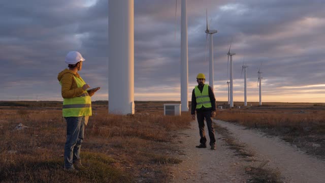可再生能源系统。慢镜头:电力维护工程师在风力涡轮机发电厂附近的田里工作，身后是阴沉的天空。两位工程师同事的商务会议。团队合作。视频素材