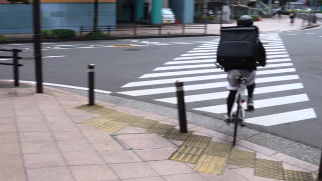 一名年轻的快递员背着黑色背包，戴着防护面具，骑着自行车在街道上送外卖。过马路。视频素材