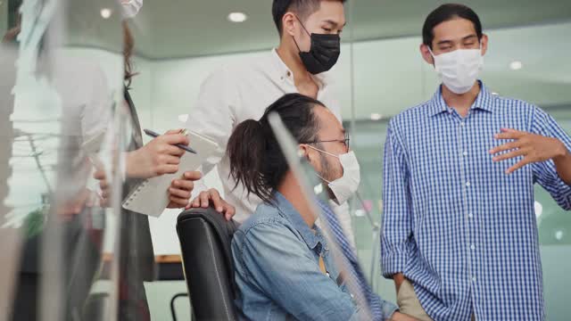 一群亚洲年轻的商务人士在办公室里用电脑工作，带着新的正常生活理念。工作场所男女应佩戴防护口罩，并保持距离，以预防冠状病毒感染。视频素材