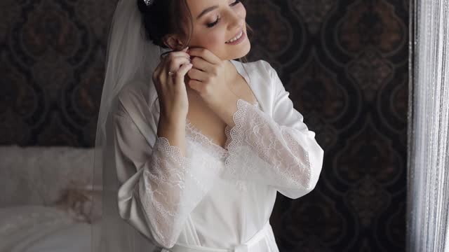 穿着闺房礼服的新娘戴上耳环。早上的婚礼准备工作。穿着睡衣和面纱的女人视频素材