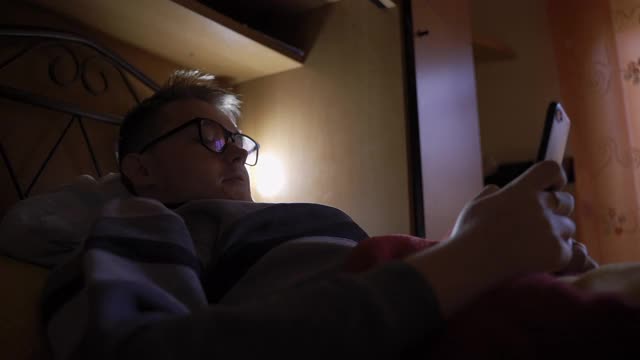 戴眼镜的年轻人晚上躺在家里的沙发上，用手机玩网络游戏。年轻玩家花时间玩3D模拟视频游戏，赌博上瘾的概念视频下载