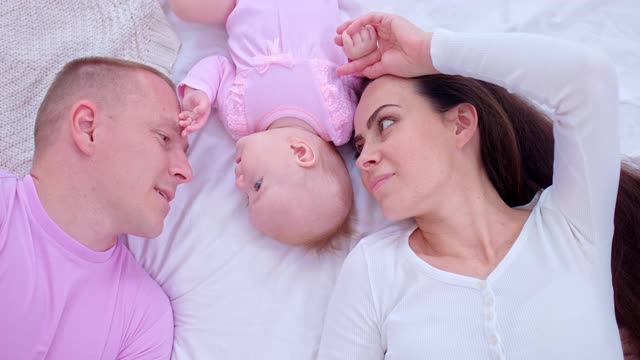全家度假时，宝宝正和爸爸妈妈躺在床上。慈爱的爸爸摸了摸宝宝的头，对她微笑。幸福的家庭。俯视图的相机。4 k, ProRes视频素材