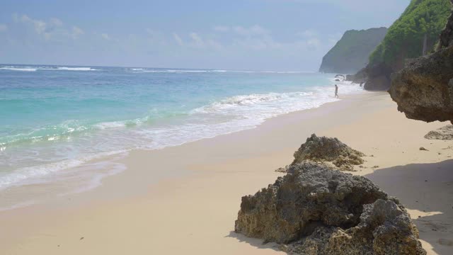 一个男人在热带海滩上走着，海浪和雾在巨大的岩石附近用慢镜头拍摄视频下载