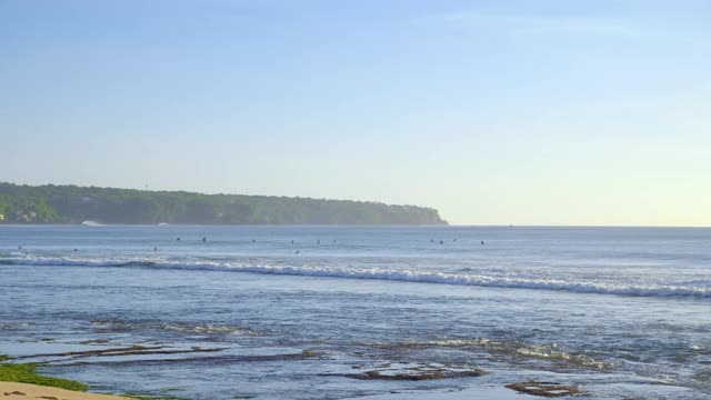 傍晚落潮时，冲浪者在岩石海滩附近的蓝色海浪中游泳视频下载