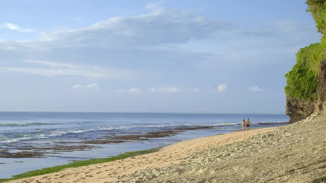 一对夫妇走在热带海滩附近的巨大岩石在低潮日落与海浪的背景视频下载