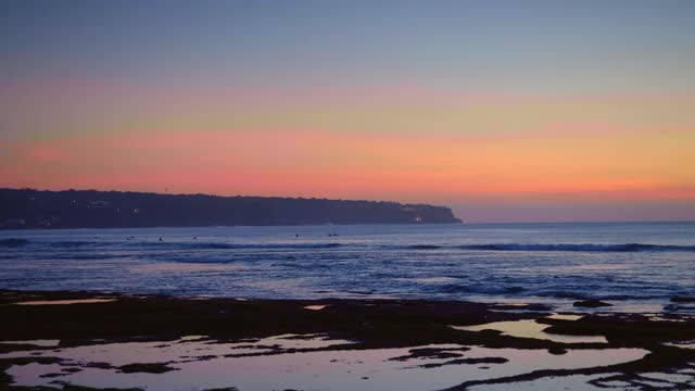 日落后的海滩和冲浪者游泳在深蓝色的水和海浪附近的岩石和悬崖视频下载