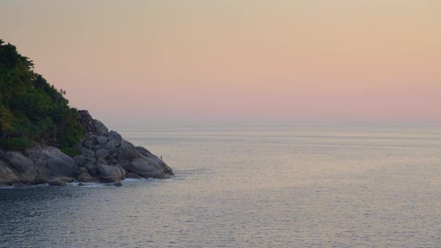 海滩与大的尖锐的石头，小山和森林在温暖的日落和渔民与rod在岩石的背景视频下载