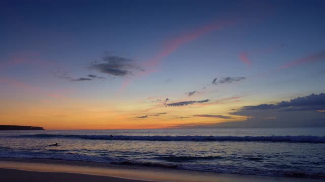日落后的海滩上，深蓝色的天空和紫色的云，冲浪者在巨浪中的剪影视频下载