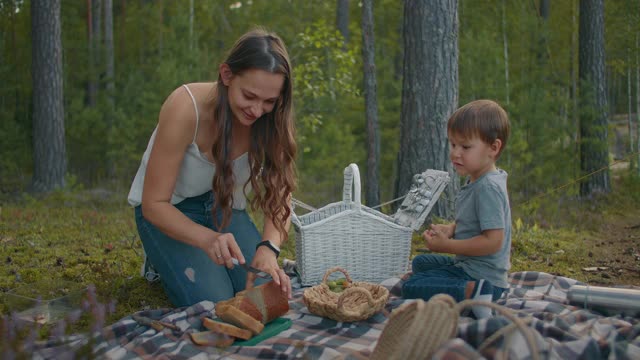 年轻女子在野餐时切面包，小男孩坐在毯子上，家人在森林里夏天休息视频下载