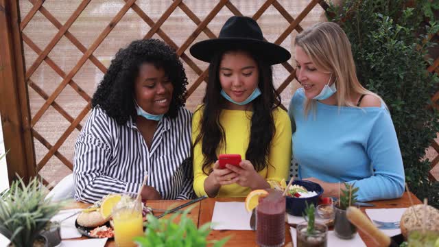 开朗的多种族女孩戴着外科口罩在早午餐酒吧看智能手机视频素材