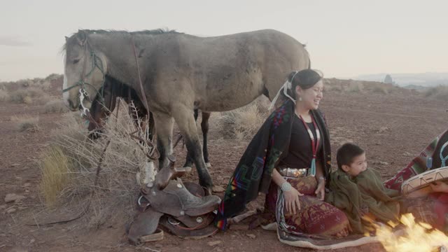 近距离拍摄的两个土著美国姐妹和他们的弟弟传统着装，纳瓦霍毯子周围他们保持温暖的篝火，他们的马和标志性的纪念碑和日落在背景视频下载