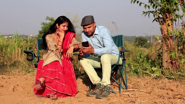 一对年轻夫妇坐在休息椅上，在农田里玩手机视频下载