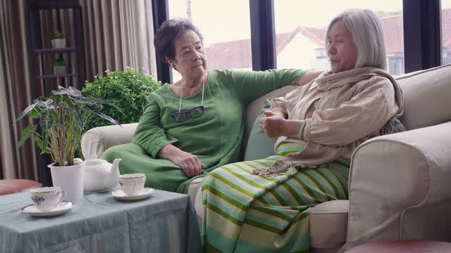亚洲资深女性朋友坐在客厅的沙发上享受下午茶，一起在家里放松和消磨时间视频下载