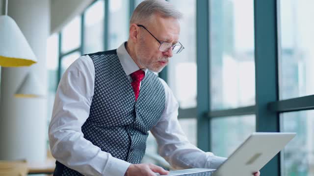 一个在电脑前工作的神经兮兮的成熟男人。戴着眼镜的严肃的资深商人站在窗边看他办公室里的笔记本电脑。视频素材