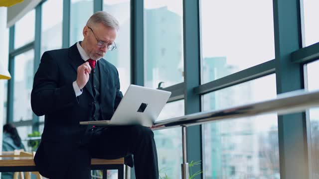 西装革履的严肃企业家。一个成熟的商人坐在桌子上，拿着笔记本电脑，若有所思地看着窗外。视频素材
