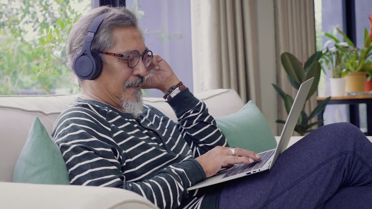 亚洲老人坐在家里客厅的沙发上，用耳机和笔记本电脑听音乐视频素材