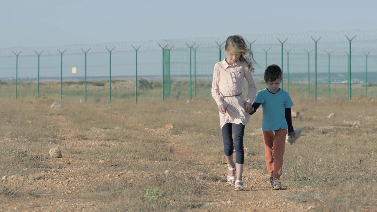 两个可怜的难民家庭的孩子，一个带着玩具的女孩和一个带着锋利的铁丝网的男孩在州边界的沙漠中行走视频下载