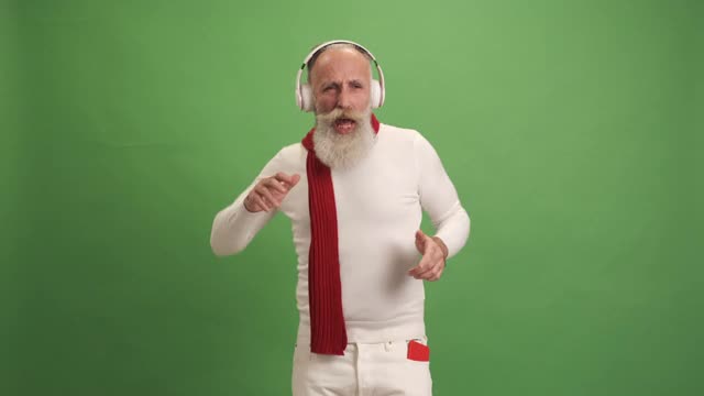 一位看起来像圣诞老人的老人用智能手机听音乐、跳舞、唱歌。在绿色的背景上。一个人。视频下载
