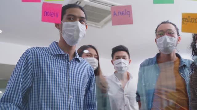 小组头脑风暴的亚洲人把便利贴贴在窗户上。团队合作的商务人士戴上口罩，可以防止新冠病毒感染，在办公室讨论和分享解决问题的想法。视频素材