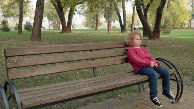 孤独的红发小孩坐在公园的长椅上，被遗弃的小孩感到心烦意乱视频下载