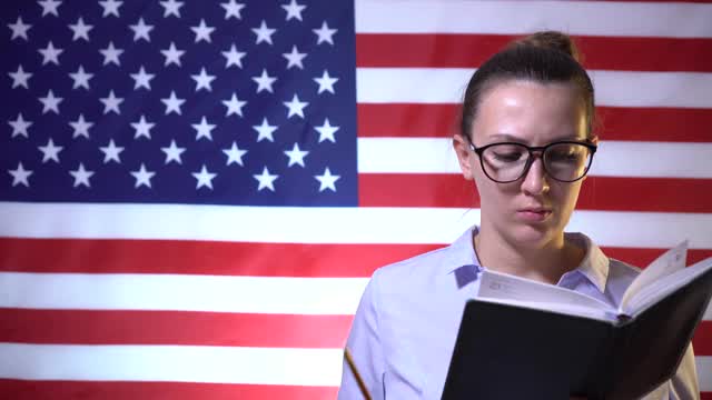 戴眼镜的女学生困惑地思考着，并在以美国国旗为背景的笔记本上写下了自己的想法。来自美国的学生，美国学生。美国教育视频下载