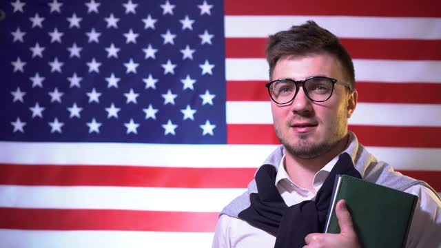 一个漂亮的学生戴着眼镜，拿着书，穿着衬衫，站在美国国旗前，微笑着，环顾四周。美国的教育。教育的概念视频下载