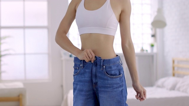 年轻苗条的女人穿着大牛仔裤在家里展示她的减肥结果视频下载