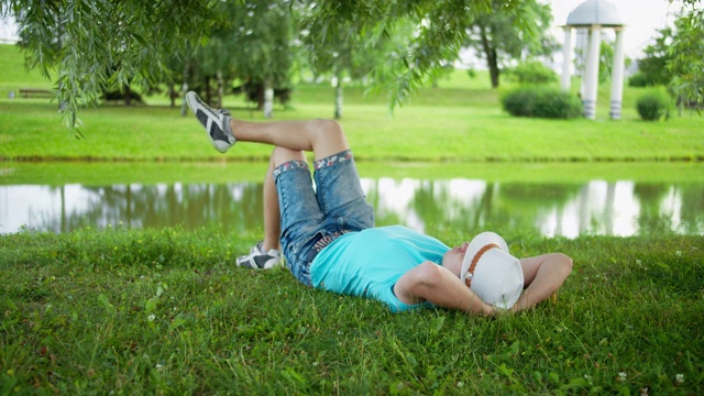 戴着帽子的快乐的年轻人躺在城市公园的一棵树下的草地上视频下载