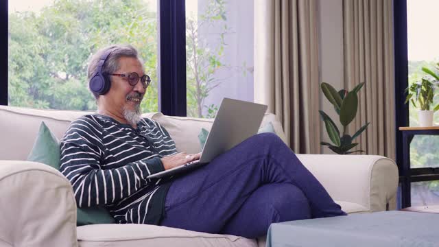 亚洲老年人坐在家里客厅的沙发上，用耳机听音乐，用笔记本电脑视频素材