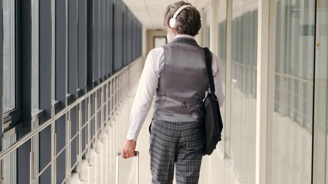 一个穿着灰色西装，带着行李箱的老嬉皮士沿着机场大厅走视频素材