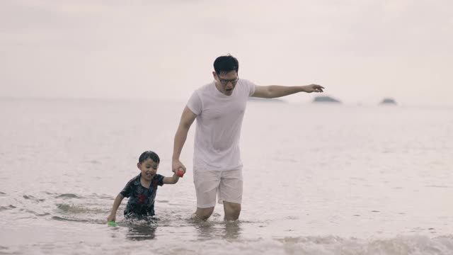 亚洲父亲和儿子一起在海滩的波浪中奔跑视频素材