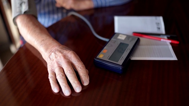 一名双手布满皱纹的老人在生病时打开数字压力装置，以测量血压和脉搏。一名男性退休人员检查高、低压数字并记录数据视频素材