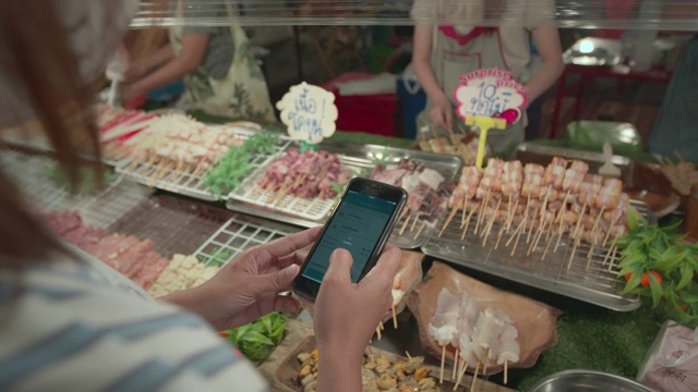 一名亚洲女子在夜市买了猪肉串、肉串和蔬菜串后，用智能手机扫描条形码付款。泰国的街头小吃。视频下载