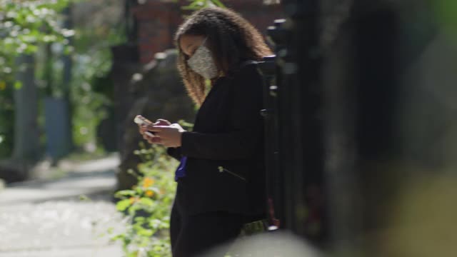 年轻的女商人靠在金属栅栏上，戴着口罩在手机上打字视频素材
