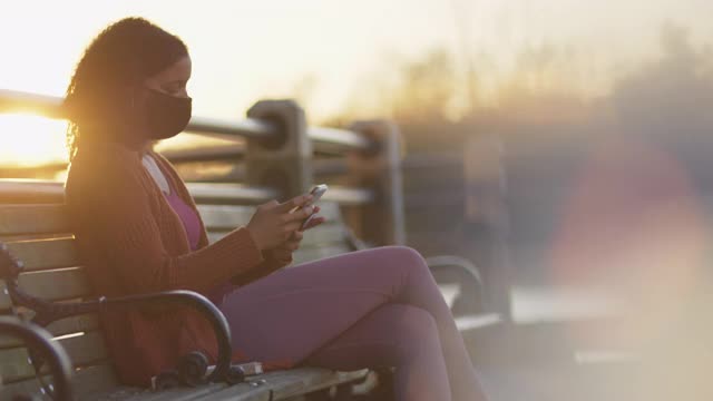 时髦的年轻女子戴着保护面罩坐在户外的长椅上，同时使用手机和听音乐视频下载