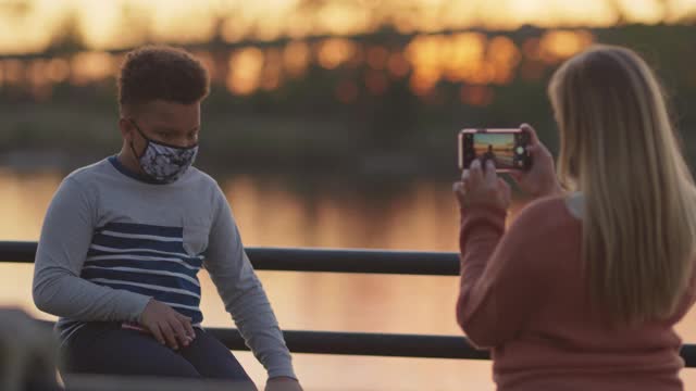 母亲给戴着防护面罩的儿子拍照，他在河边摆姿势视频素材