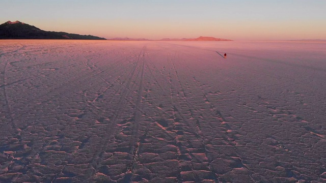 一个人走在博纳维尔盐滩上的航拍视频剪辑，与日落的怪异的粉红色辉光和汽车在高速公路上行驶留下的轮胎痕迹视频素材