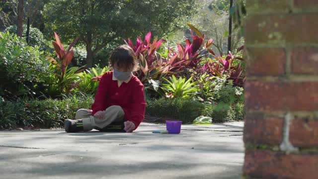 可爱的小男孩戴着面具坐在人行道上用粉笔画画视频素材