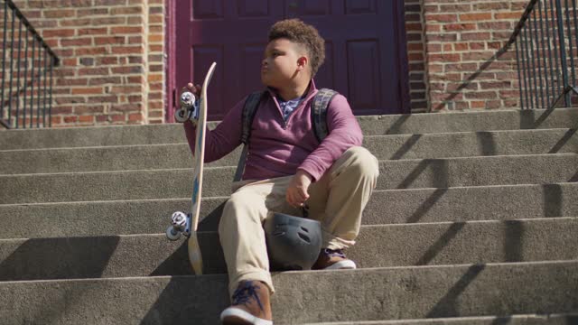 一个自信的小男孩拿着滑板和头盔坐在楼梯上看着摄像机视频下载