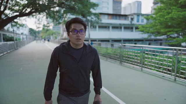 在泰国曼谷市中心商业区，一名戴眼镜的亚洲青年男子在跑步前休息喝水的慢动作视频素材