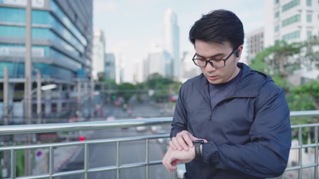 在泰国曼谷市中心商业区跑步前，一名年轻的亚洲男子戴着眼镜和智能手表休息的慢动作视频素材