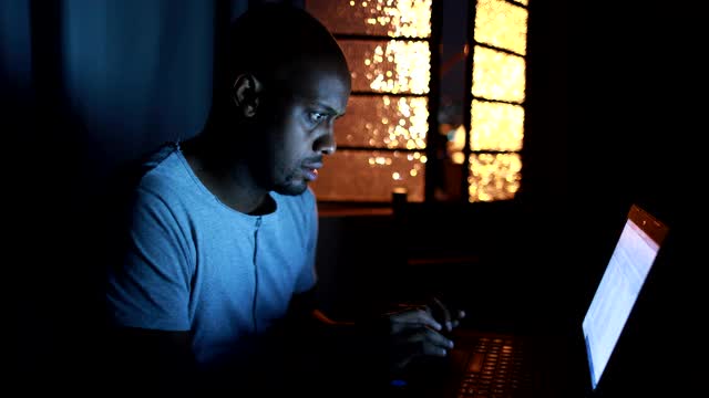 非洲人在黑暗中看着笔记本电脑屏幕，一个人在电脑前工作到很晚视频素材