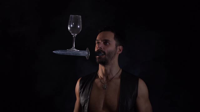 一个男人嘴里叼着一把刀，摇晃着，保持着杯子的平衡。独特的马戏团的技能。视频下载